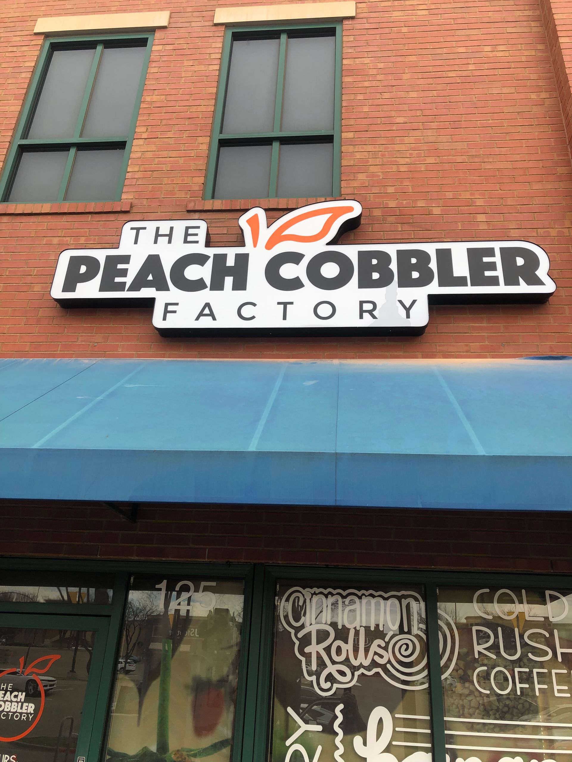 Storefront sign for “The Peach Cobbler Factory” on a brick building with a blue awning.