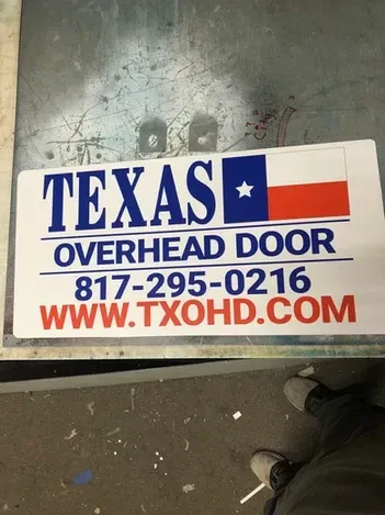 Texas Overhead Door sign on a floor, with blue, red, and white text and phone number 817-295-0216.
