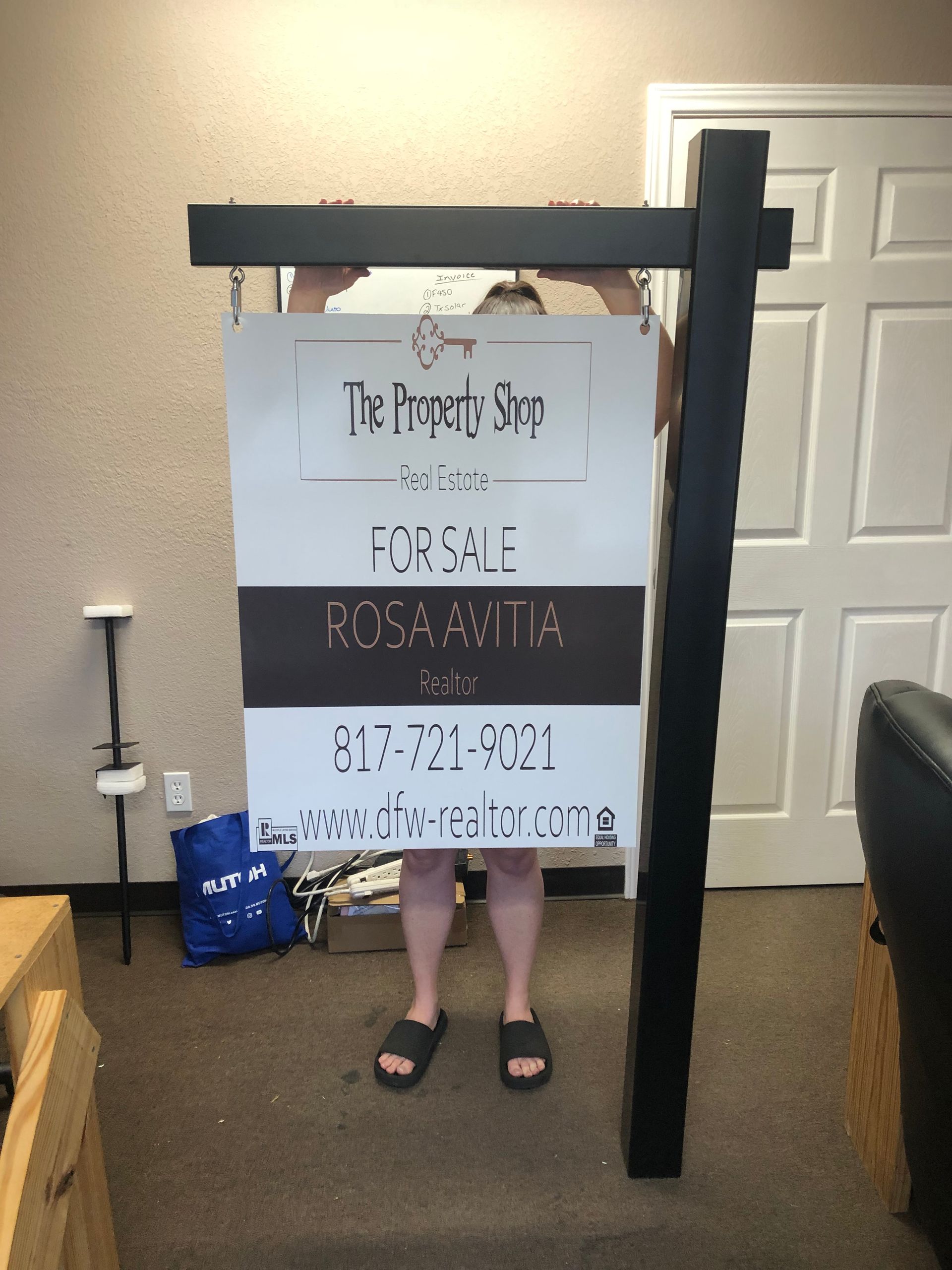 Person holding a large sign in front of a house, with a black post and a blue bag nearby.