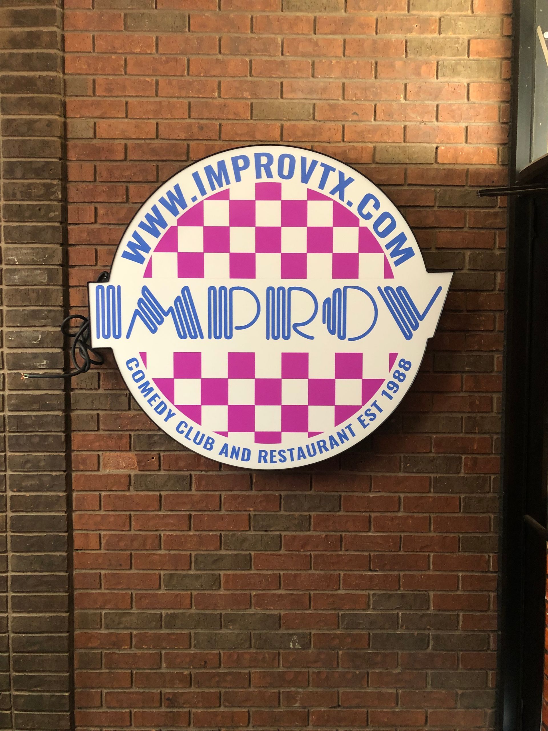 Circular IMPROV sign on a brick wall, with blue text and pink checkerboard accents.