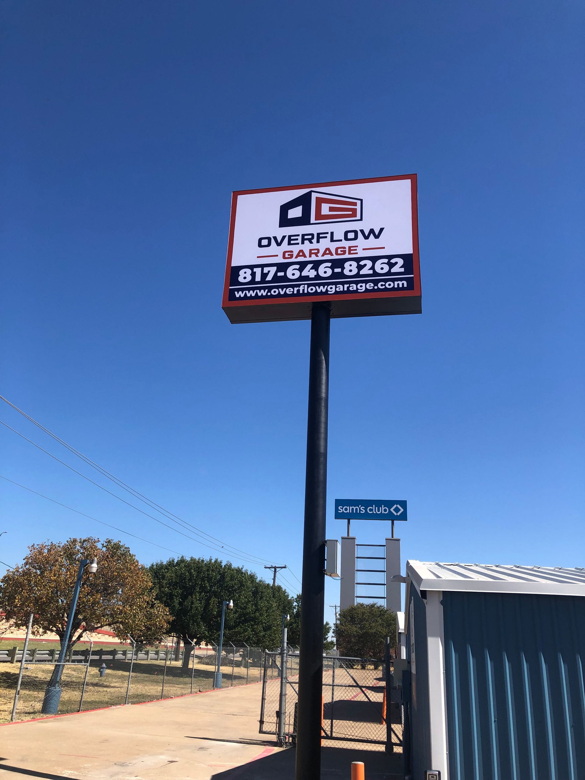 Outdoor sign for “Overflow” against a clear blue sky beside a fenced lot and shed
