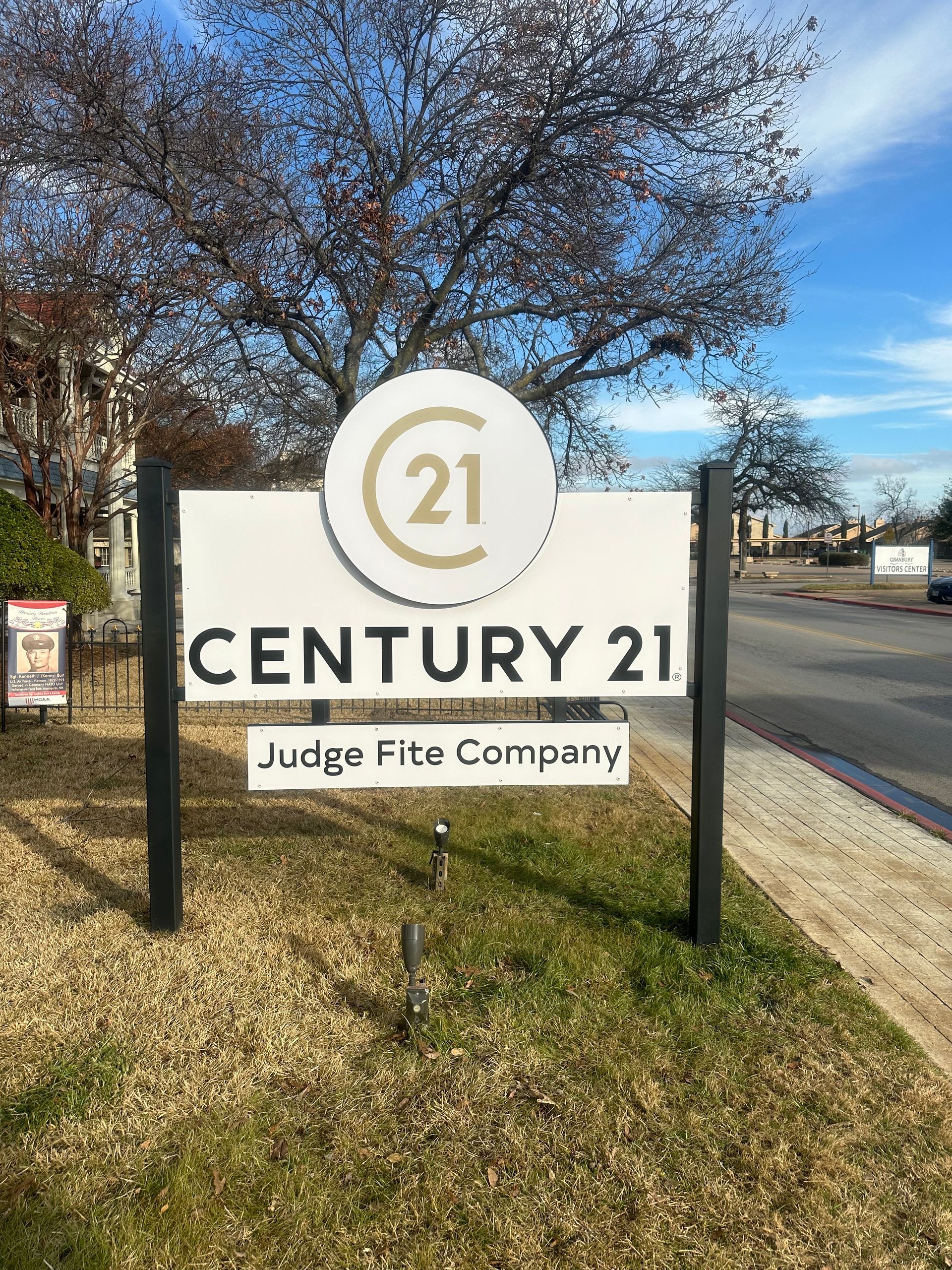 Century 21 Judge Fite Company roadside sign on a grassy corner, with trees and blue sky.