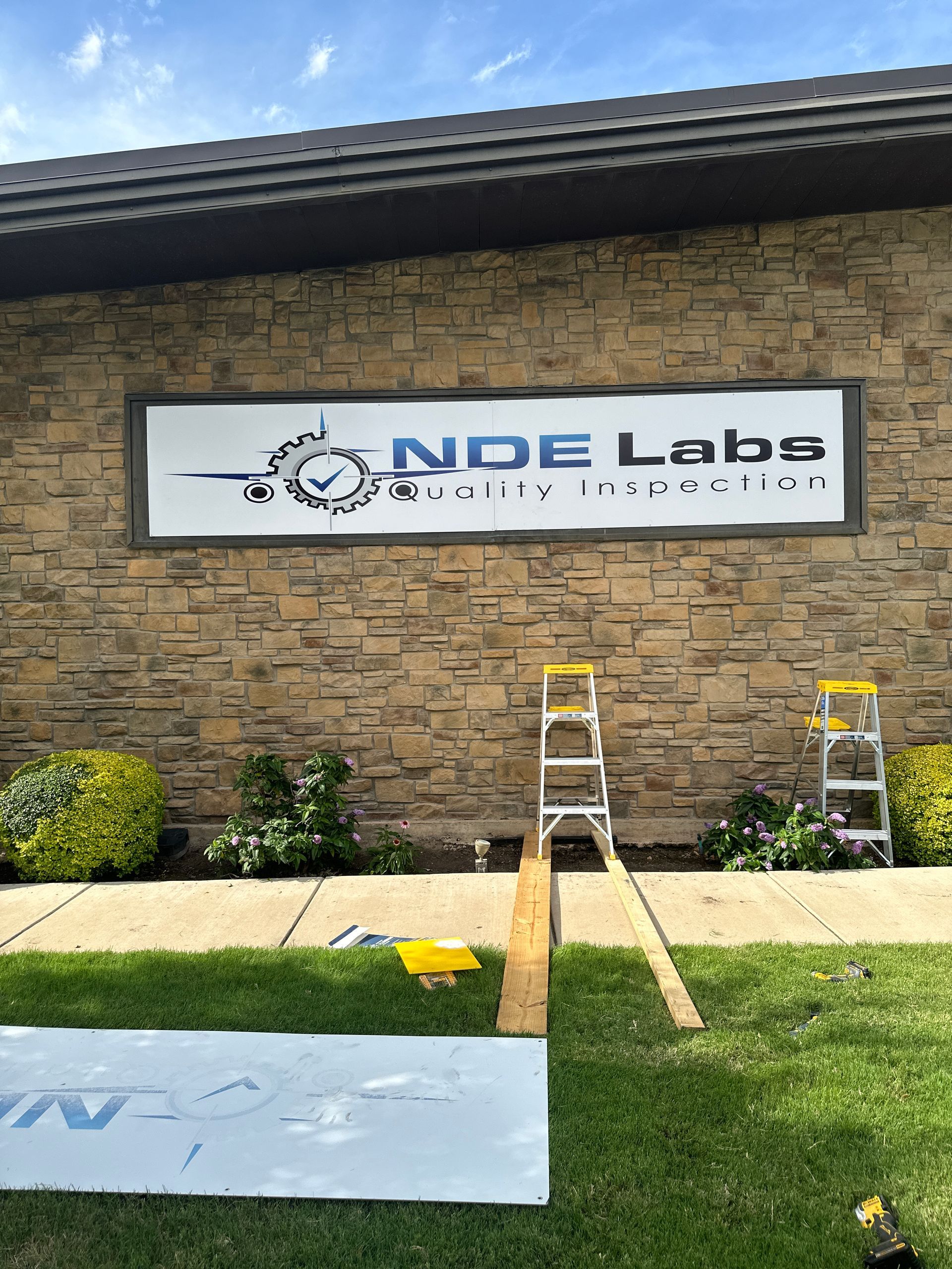 NDE Labs storefront sign on stone wall with ladders and a grassy strip in front