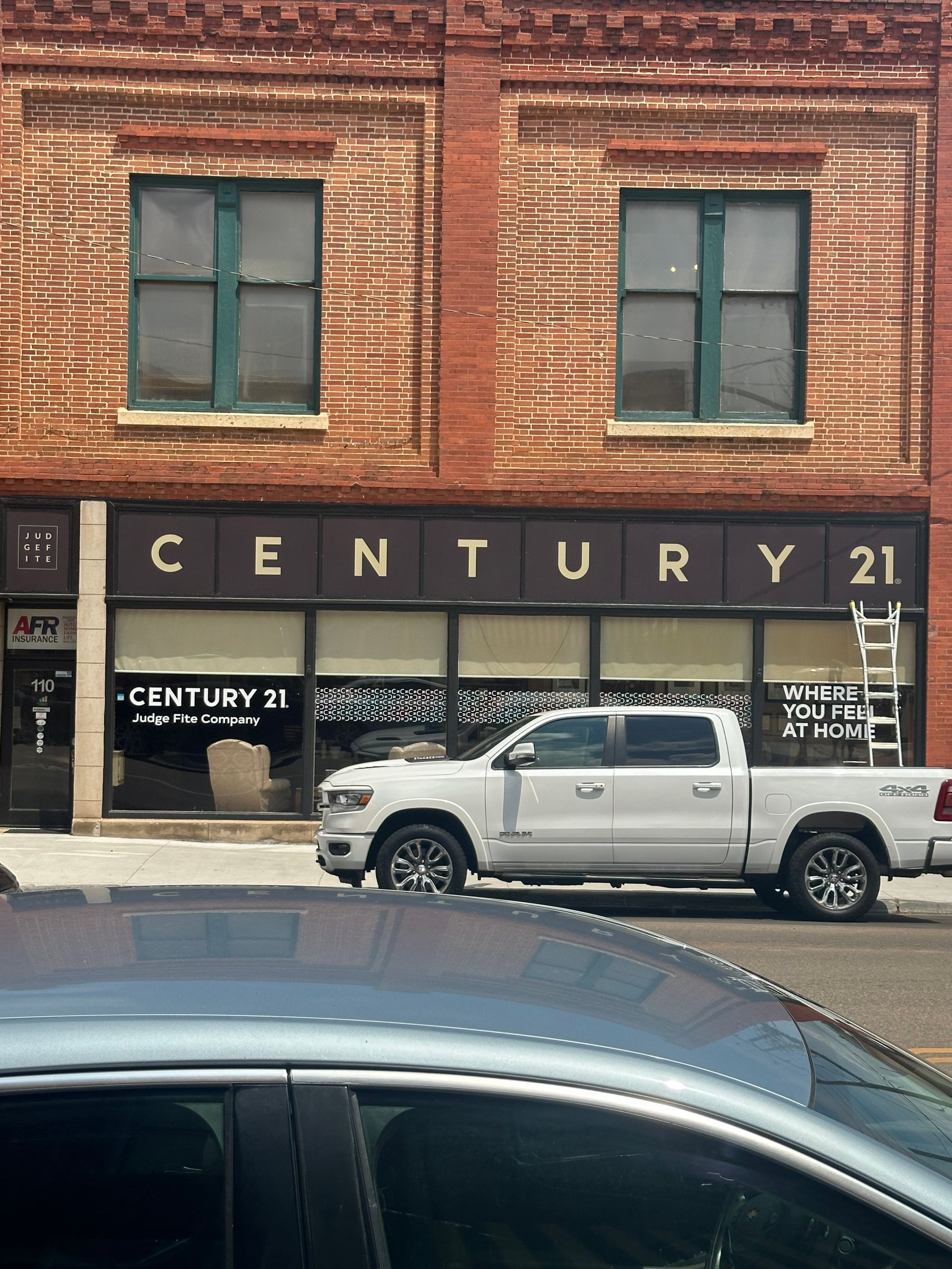 Century 21 real estate storefront in a brick building with a white pickup truck parked outside.