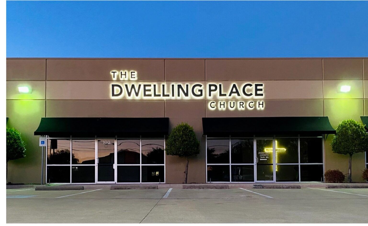 Exterior of The Dwelling Place church storefront at dusk, with lit windows and parking lot