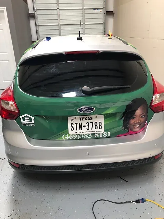 Silver hatchback parked in garage, rear view with green wrap, Texas plate, and photo decal on the back.