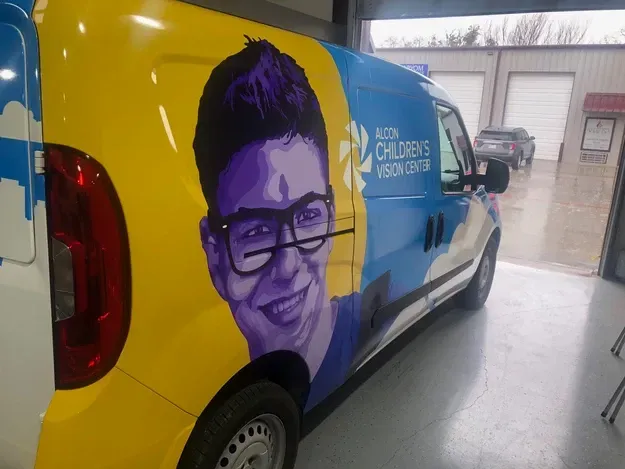 Brightly wrapped service van with a large smiling portrait and blue-yellow branding in a garage area