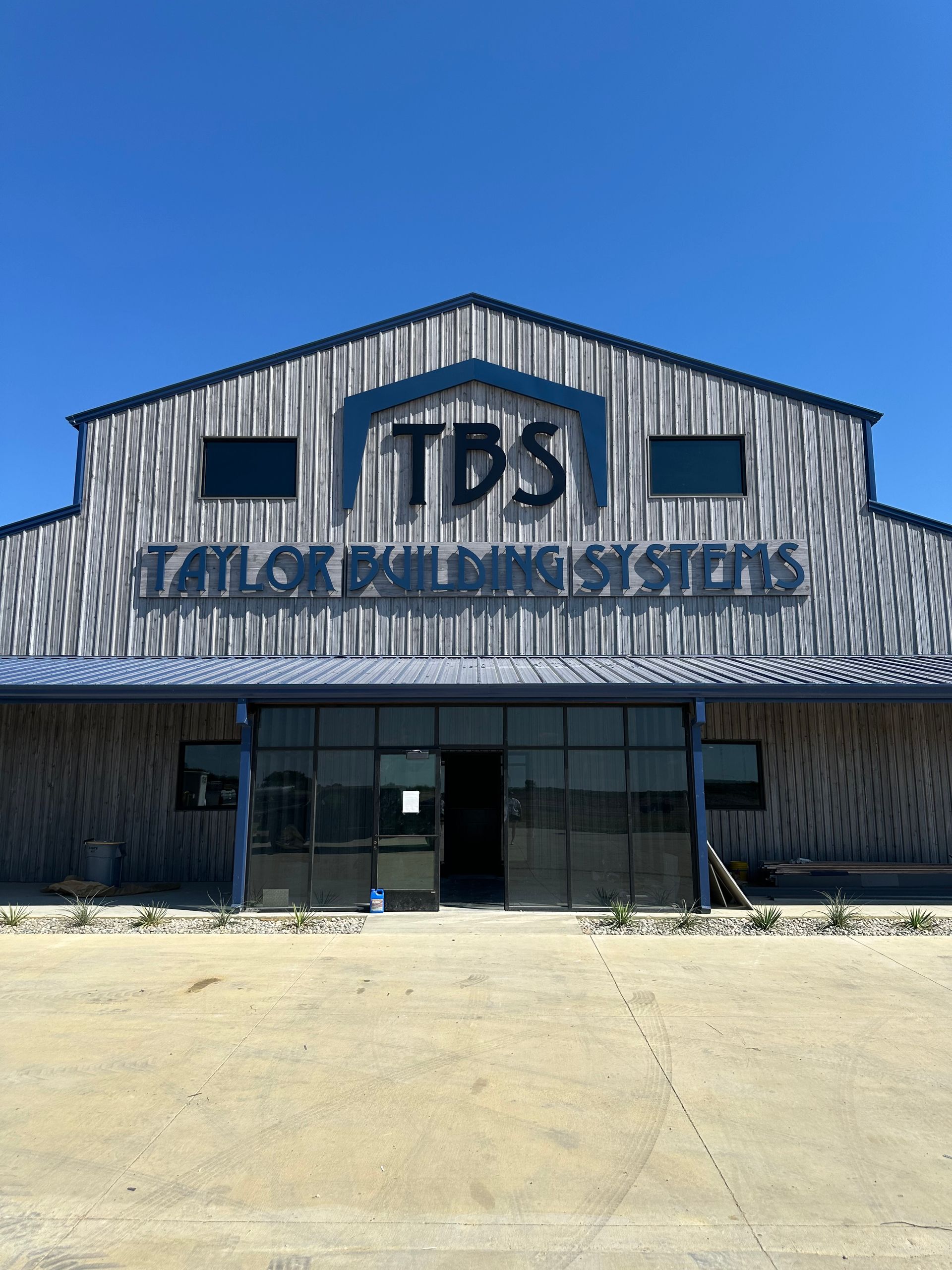 Front view of a TBS building with blue siding, glass entrance, and a large concrete lot under a clear sky