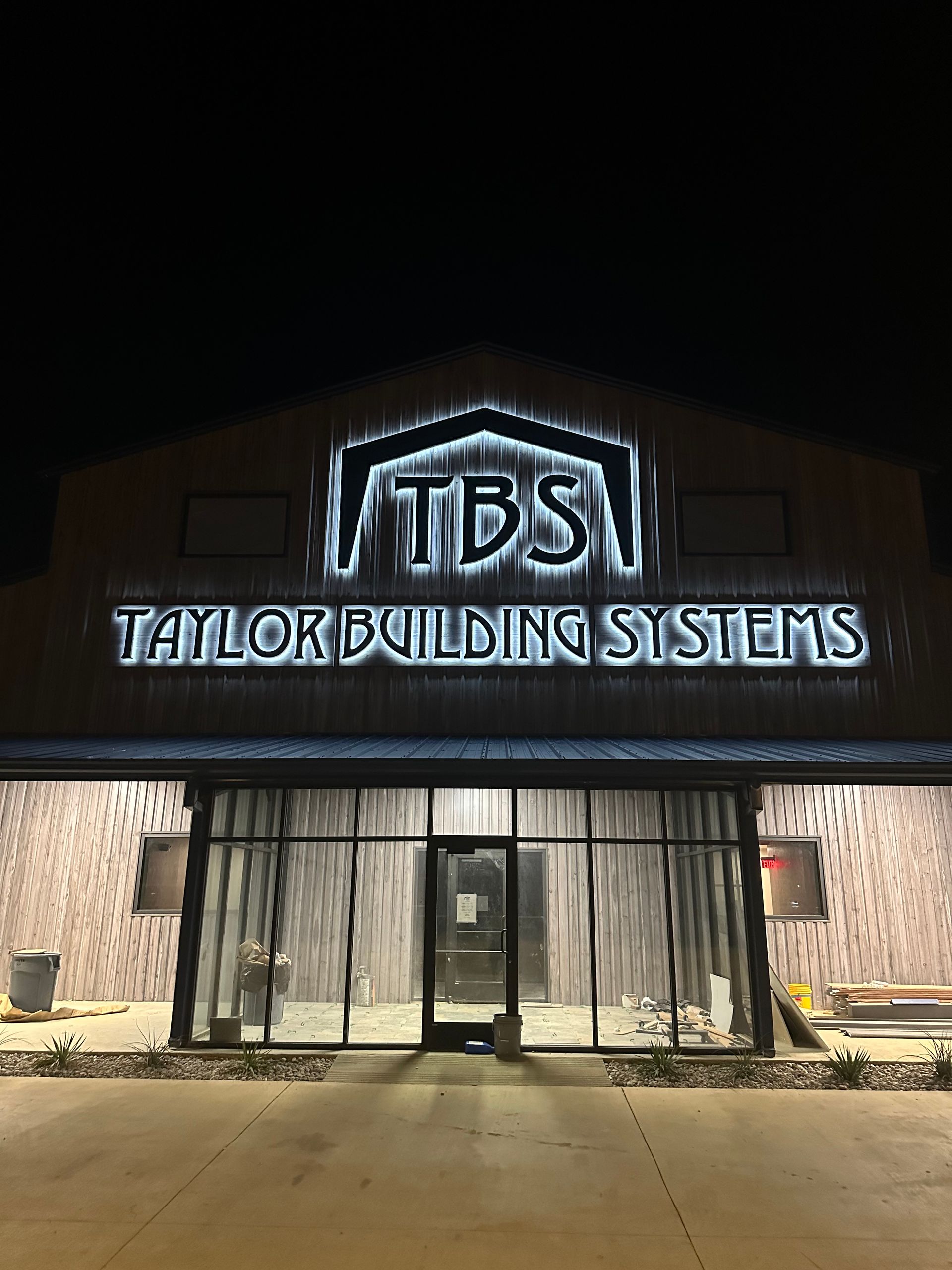 Lit-up Tailor Building Systems storefront at night with TBS sign and glass entrance