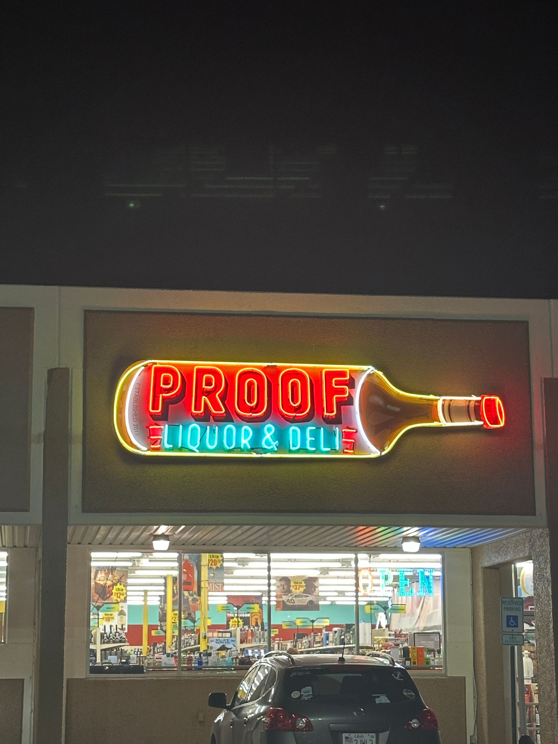 Neon “Proof Liquor & Deli” sign above a convenience store at night, with parked cars out front