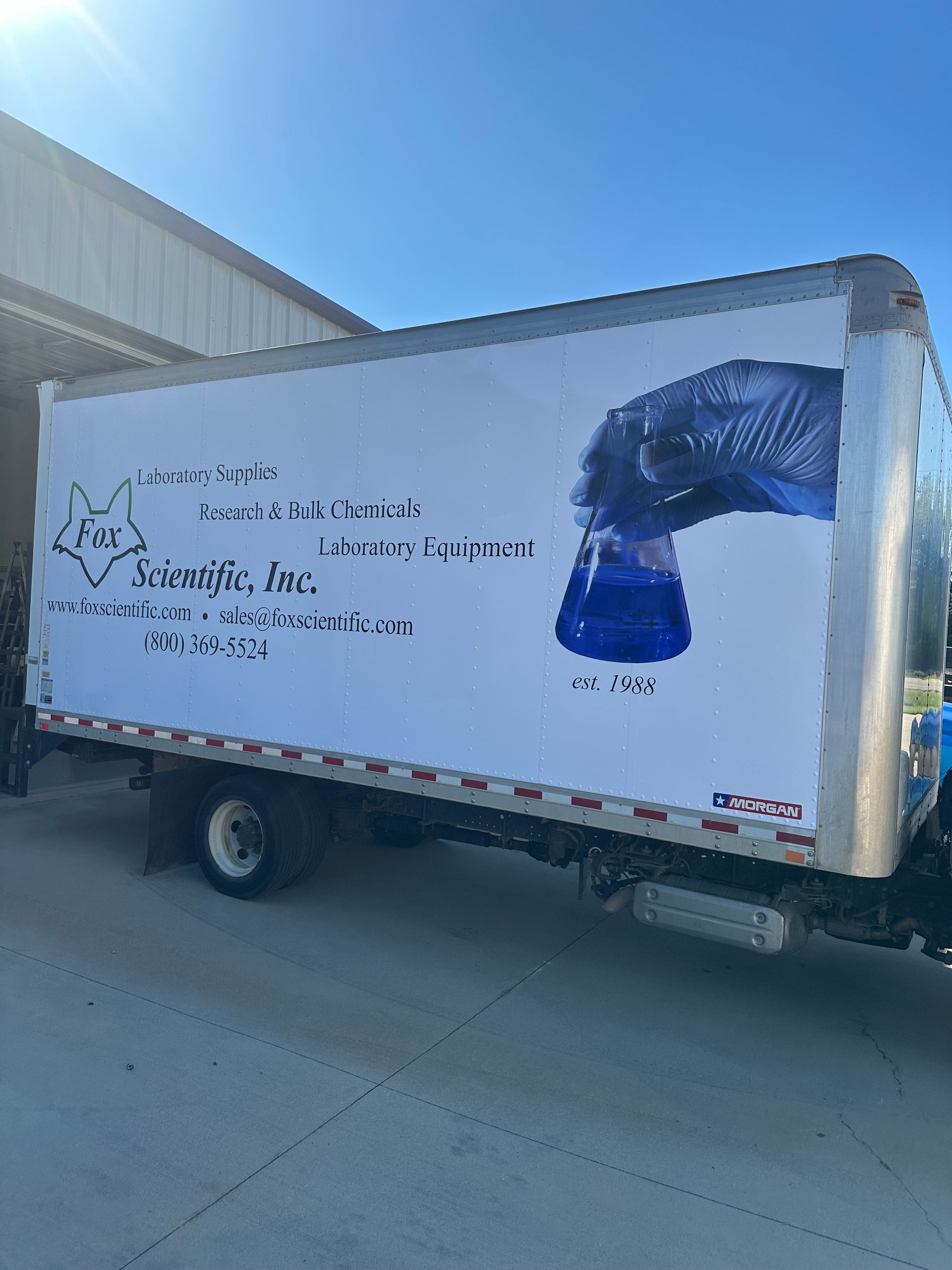 White box truck parked by a warehouse, with blue graphics and text on its side.