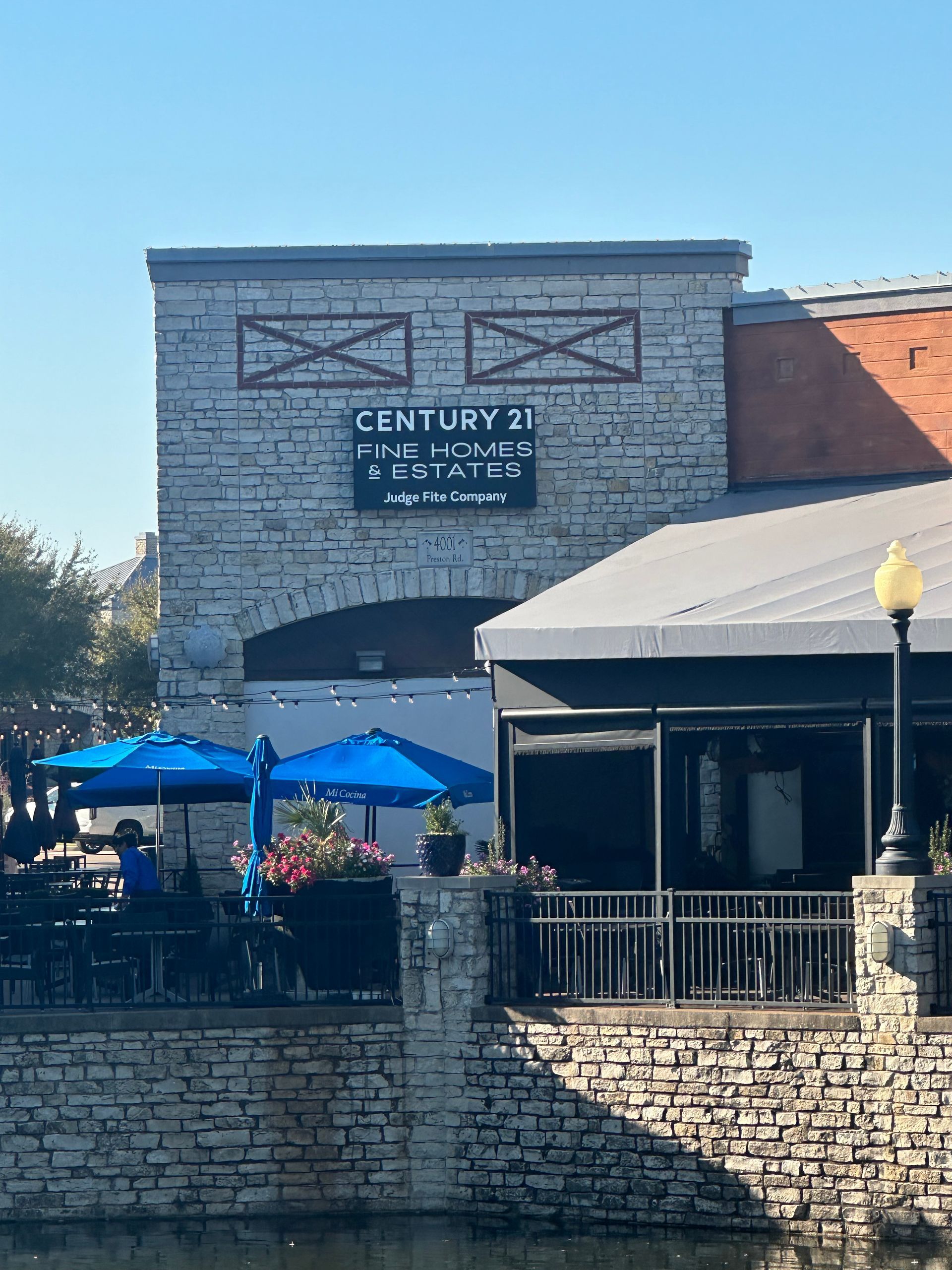 Stone building with a Century 21 sign, blue umbrellas, and a patio under a clear sky.