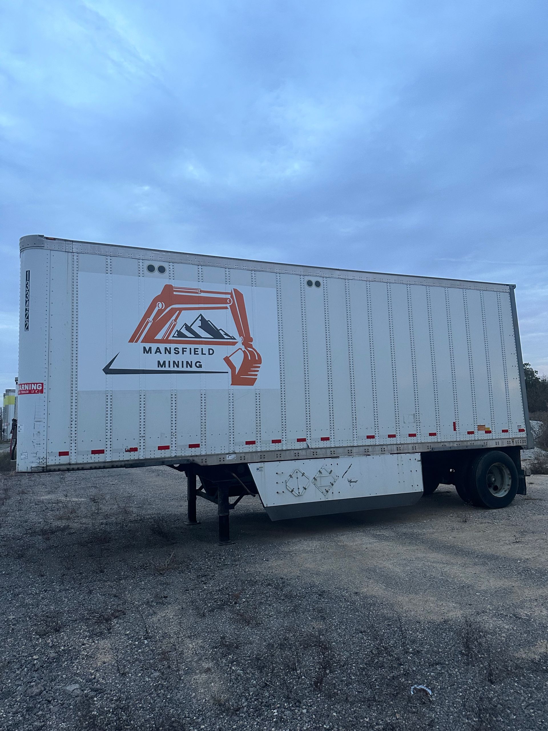 White enclosed trailer with orange logo parked on gravel under a cloudy sky.