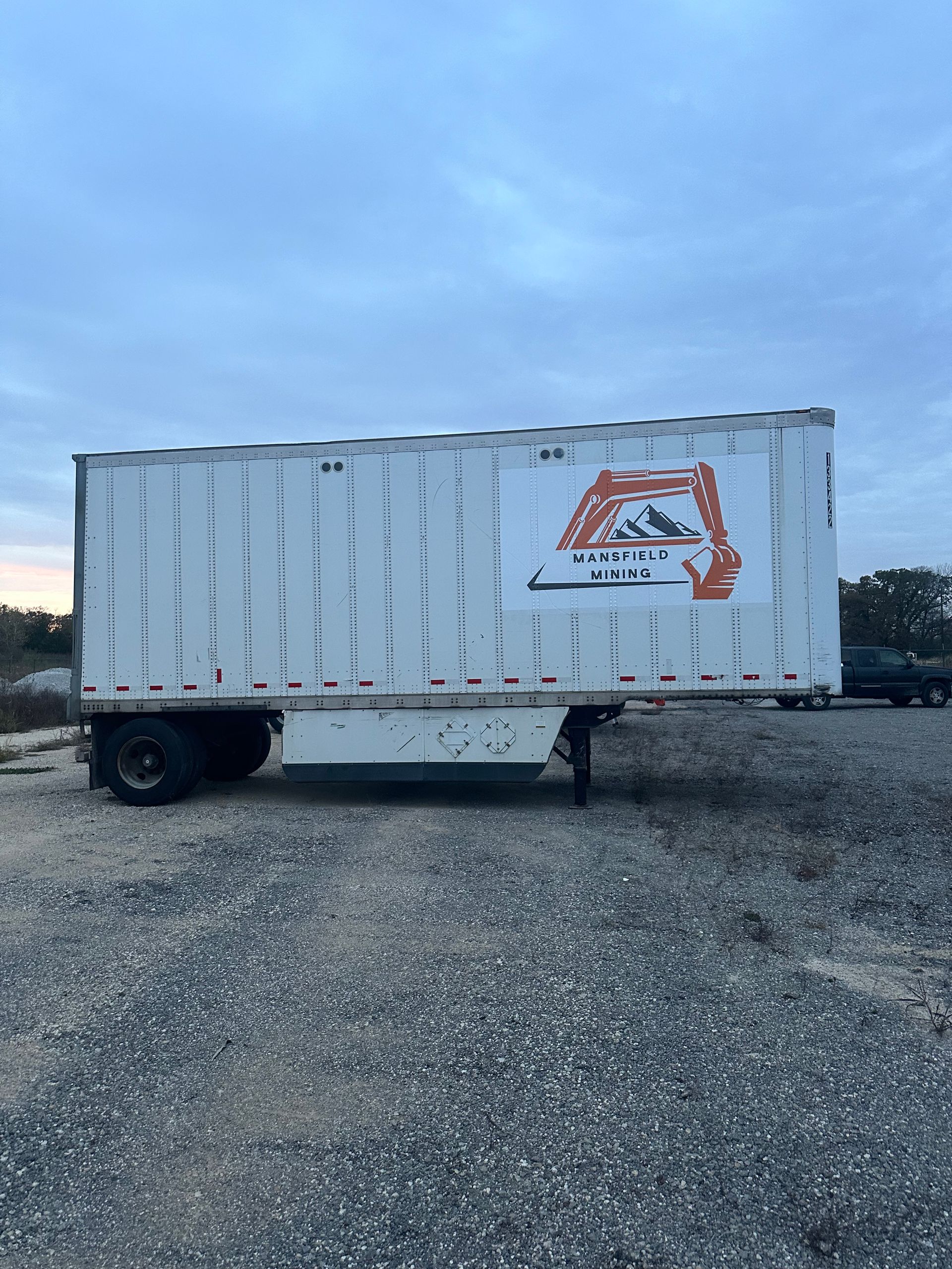 White box truck trailer on gravel, with an orange logo on the side, parked outdoors at dusk.