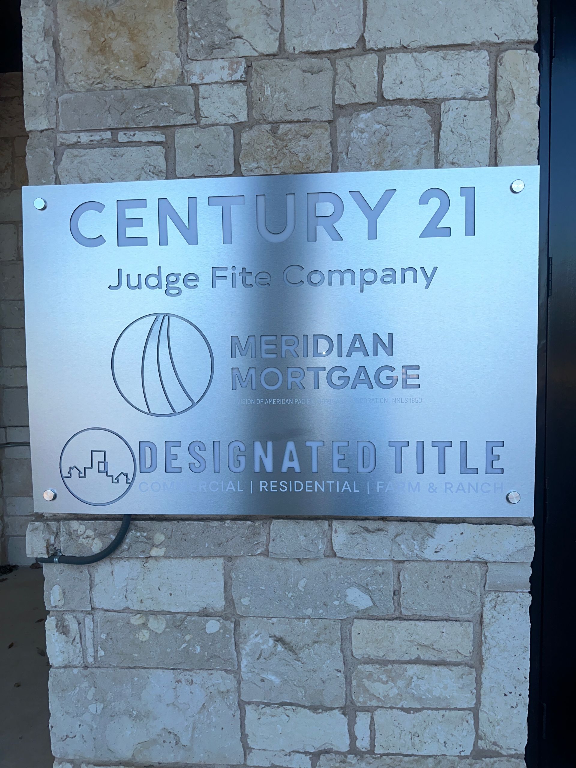 Century 21 real estate sign mounted on a stone wall, showing office and agent names.
