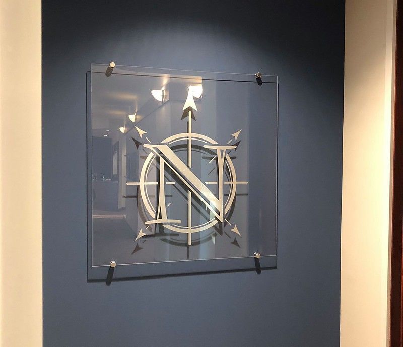 A clear square sign mounted on a dark blue wall displays a silver compass rose graphic with a large letter N in the center.