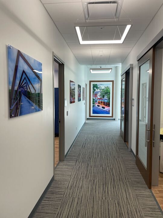 An office hallway featuring grey carpet, white walls adorned with framed landscape photos, and glass-paneled office doors.