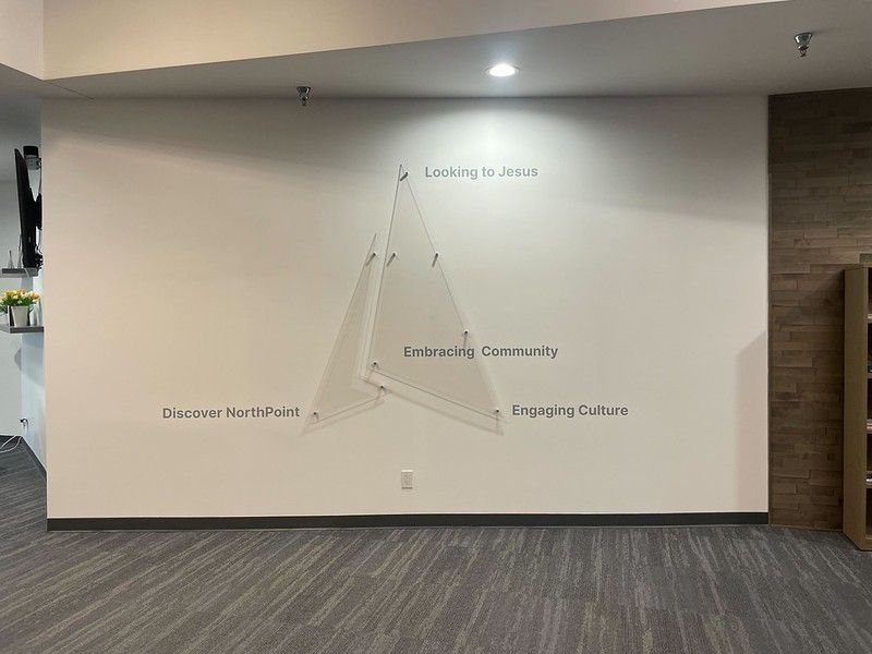 A white wall features a minimalist, triangular glass structure with text reading 