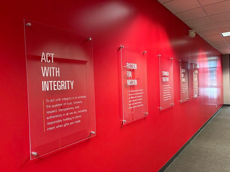A red wall lined with clear, acrylic signs displaying corporate values and mission statements in a hallway.