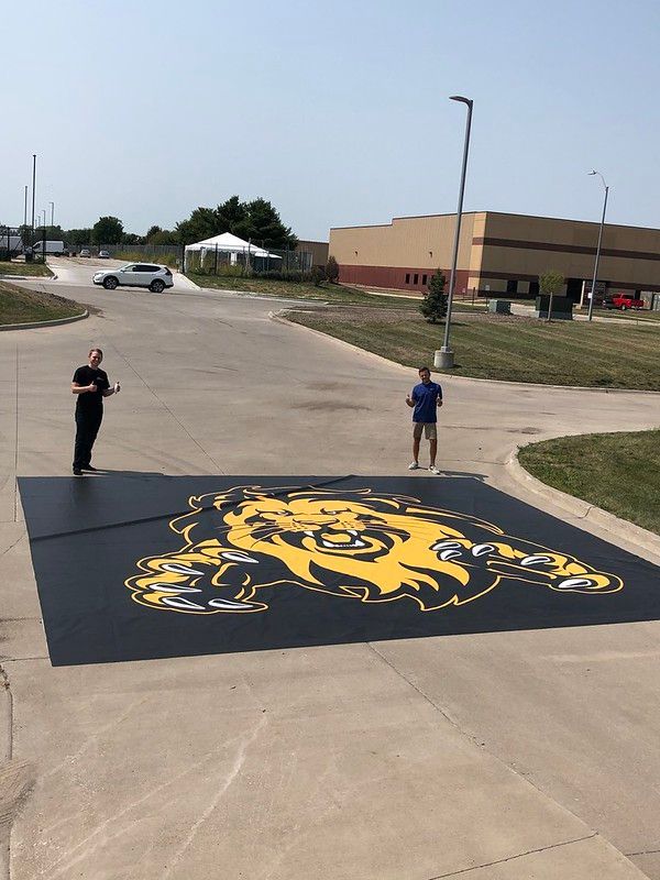 Two people stand on a parking lot beside a large black-and-yellow lion decal applied to the ground.