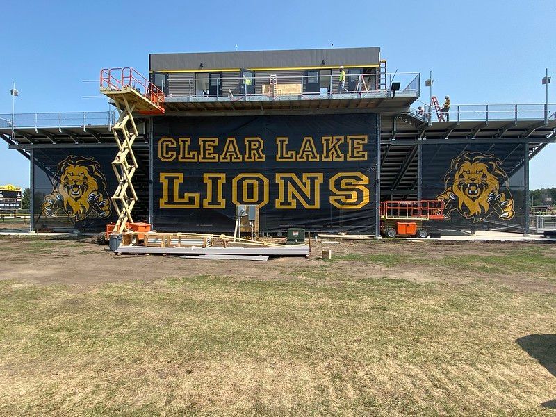 Clear Lake Lions stadium bleachers with a large team logo banner, undergoing construction with scissor lifts in front.