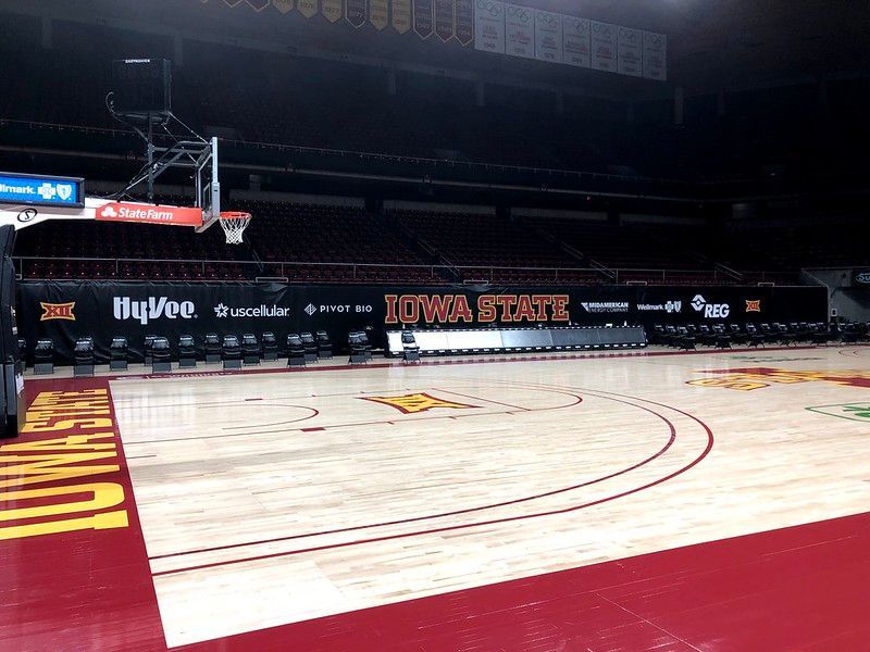 The Iowa State basketball court features a prominent logo, maroon painted borders, and a Hy-Vee advertisement on the wall.