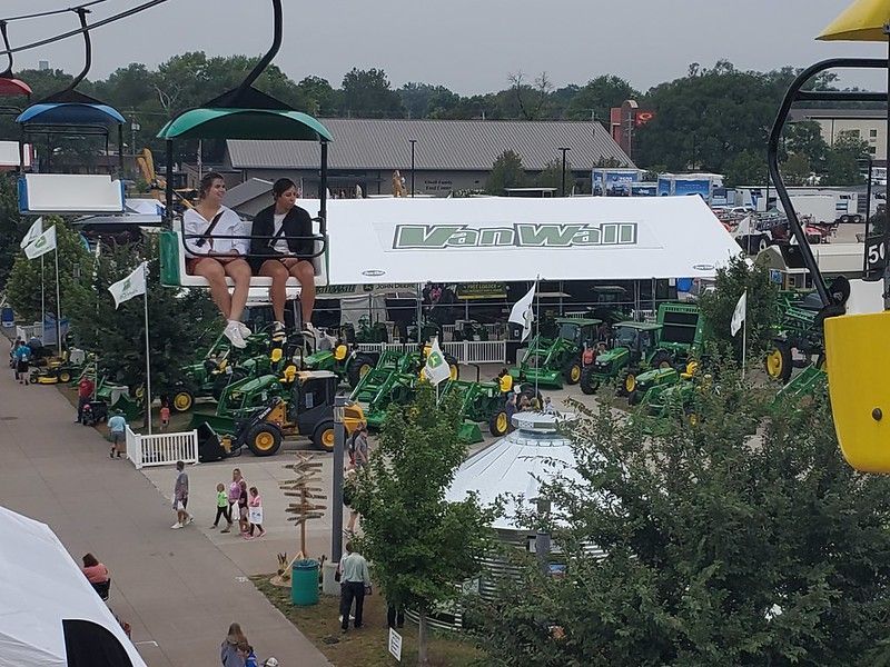 People ride a ski lift over an outdoor display of John Deere agricultural machinery at a fairgrounds.