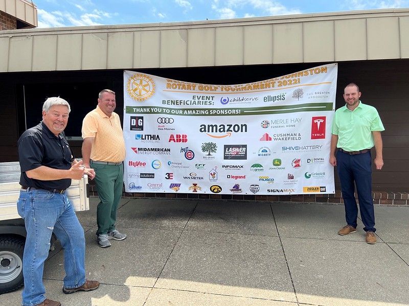 Three people stand in front of a Rotary Golf Tournament sponsor banner outdoors on a sunny day.