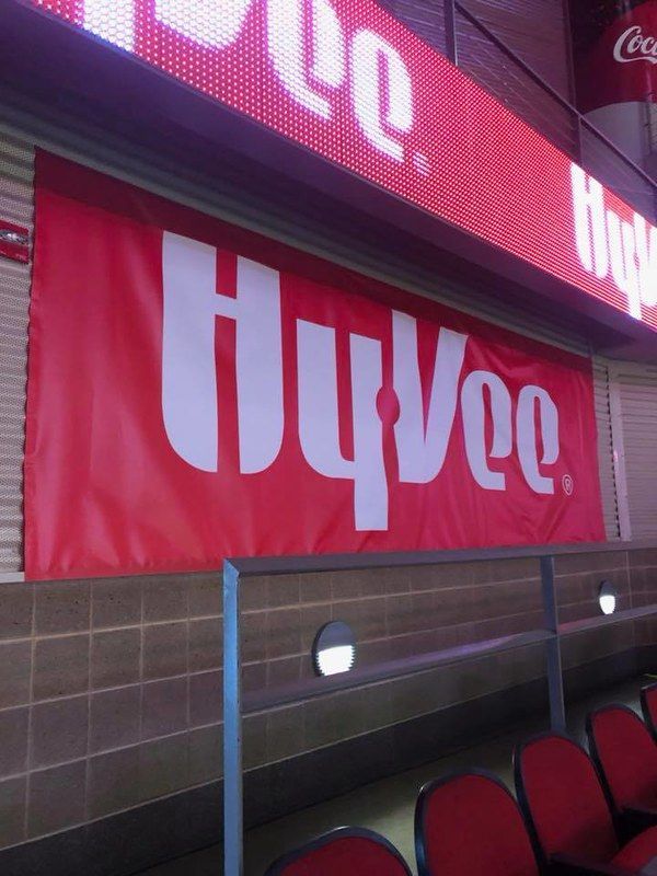 A large red Hy-Vee banner hanging inside a stadium seating area with a digital sign visible above it.
