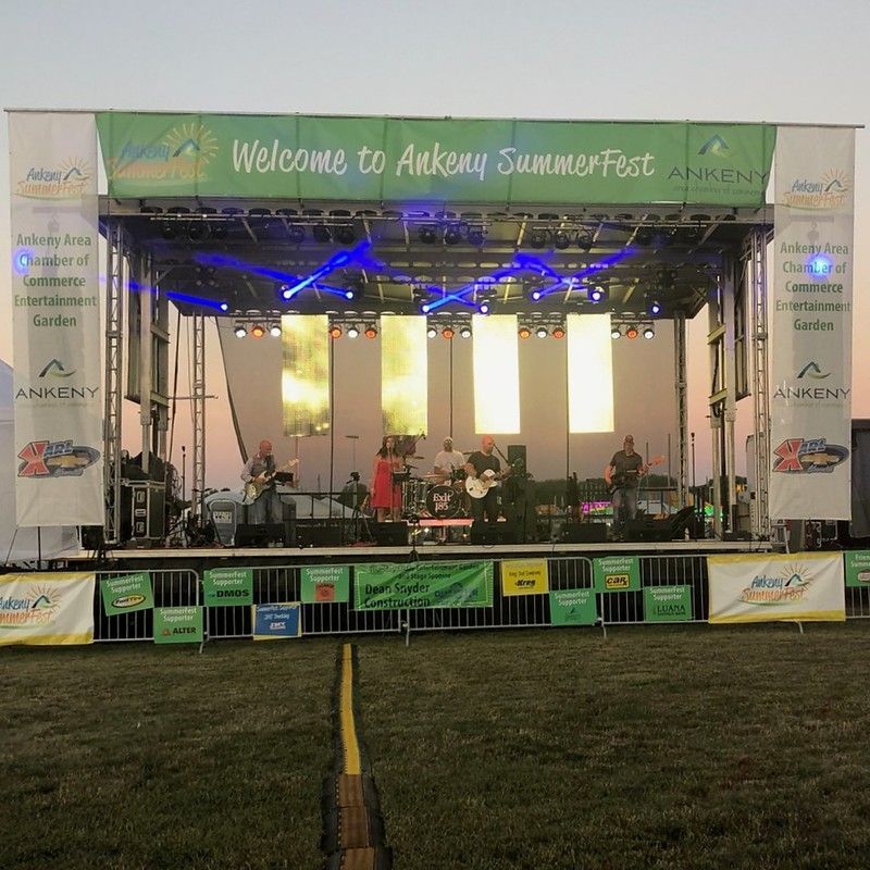 A band performs on a stage during Ankeny SummerFest, with large banners, lighting, and a field in the foreground.