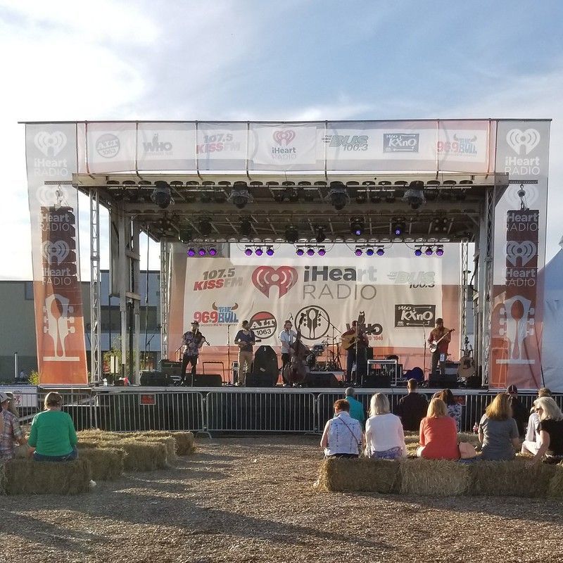 A live band plays on an outdoor stage with iHeartRadio banners while an audience sits on hay bales in front.