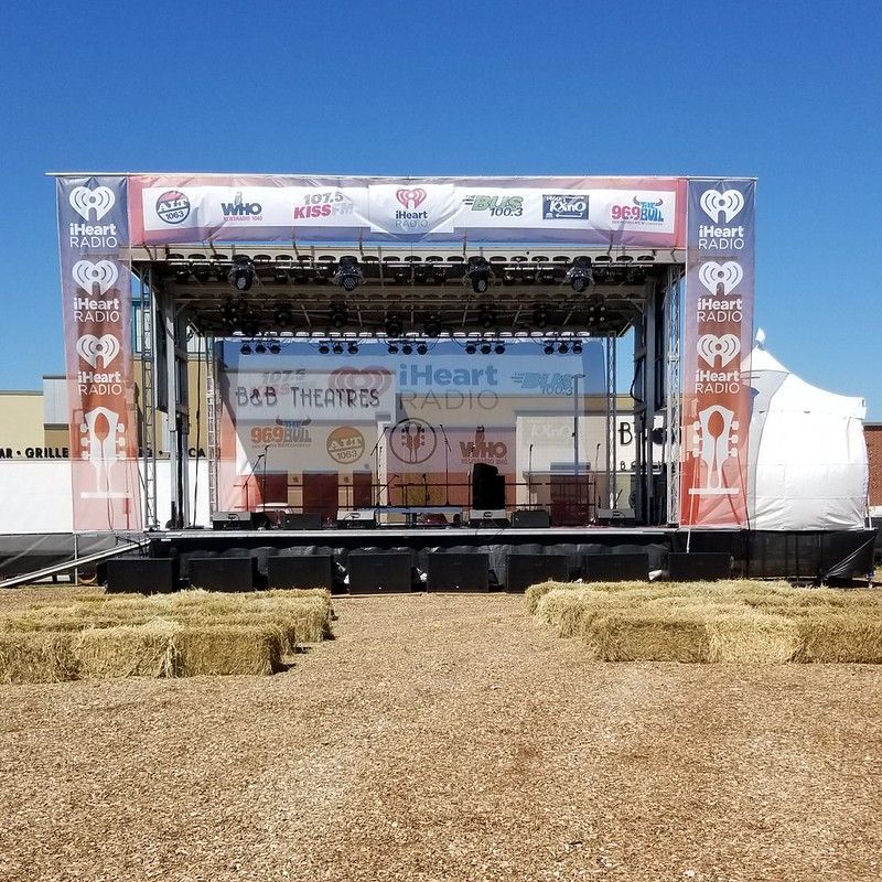 An outdoor iHeartRadio stage set up with hay bale seating on a dirt ground under a clear blue sky.