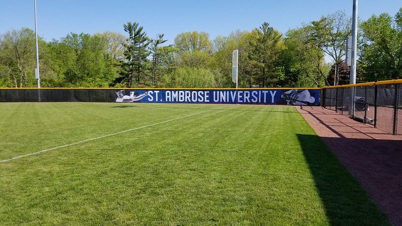 A St. Ambrose University branded fence lines a lush green baseball field under a clear blue sky.