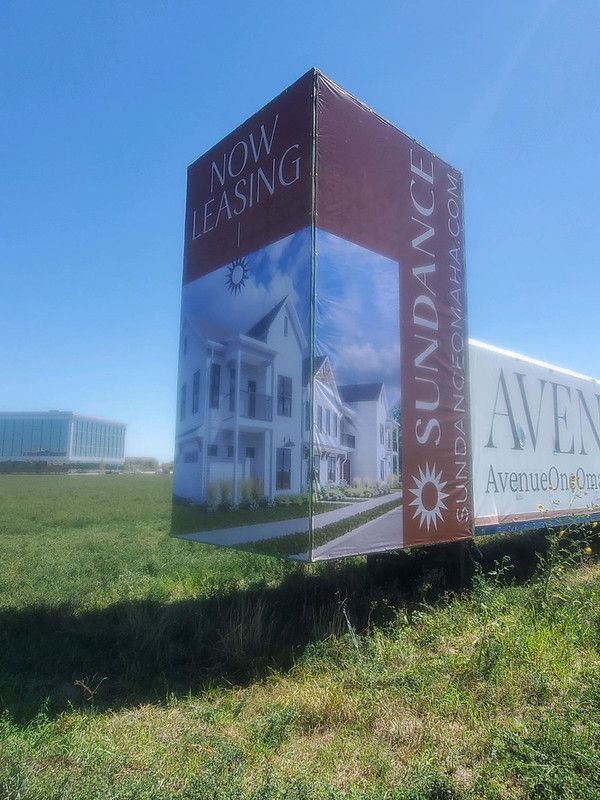 A billboard advertising Sundance apartments, displaying a picture of a house, against a bright blue sky and grassy field.