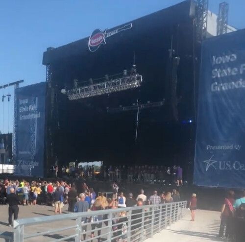 An outdoor concert stage with large blue banners at the Iowa State Fair, with a crowd of attendees gathered in front.