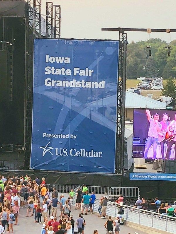 A large blue Iowa State Fair Grandstand banner at an outdoor venue, with a crowd of people in front and a concert screen.