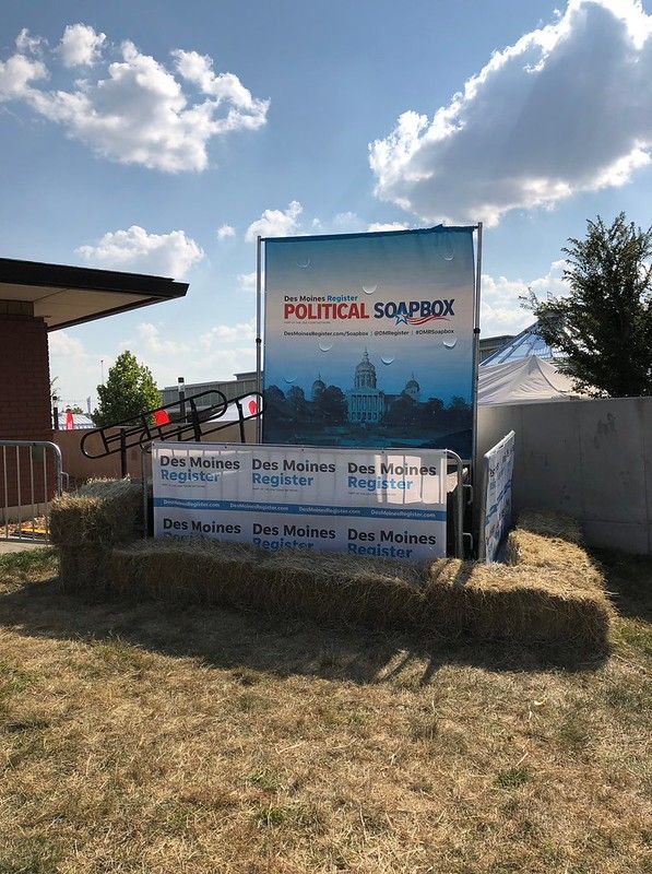 A Des Moines Register Political Soapbox platform, built with hay bales, sits outdoors under a blue sky with clouds.