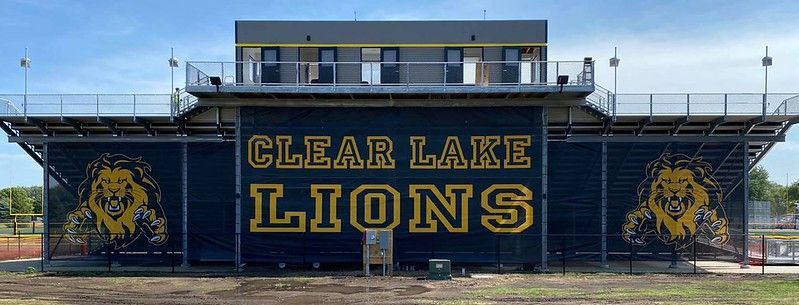 Clear Lake Lions football stadium bleachers with team logos on a black background under a blue sky.