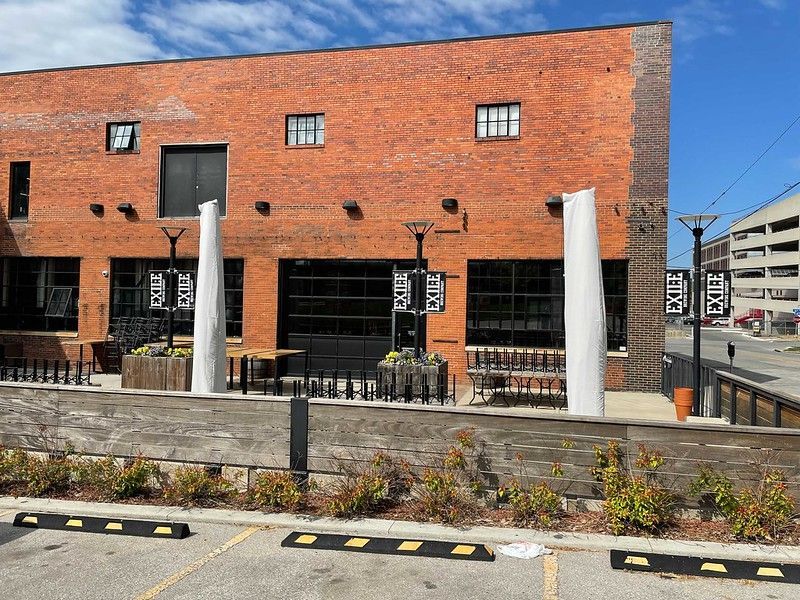 A brick building with outdoor patio seating under large white umbrellas, next to a parking lot with yellow-marked curbs.