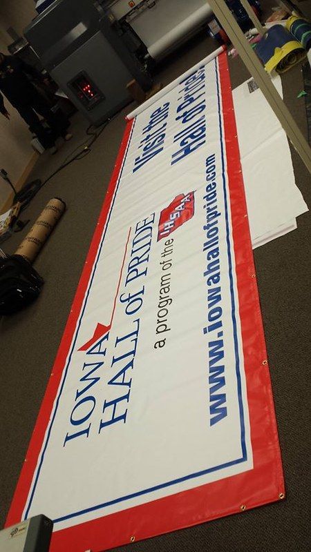 A long, white vinyl banner for the Iowa Hall of Pride, featuring blue and red text, lying on an indoor floor.
