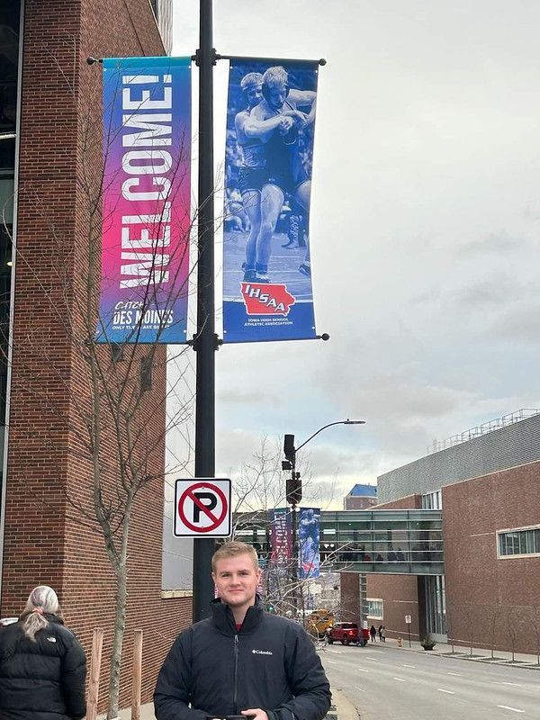A man in a black jacket stands on a city sidewalk beneath blue and pink event banners featuring a wrestler.
