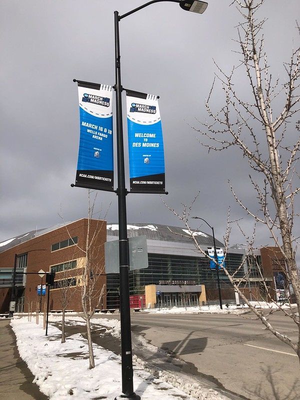 Two banners hang from a light pole in front of the Wells Fargo Arena in Des Moines, with snow on the ground.