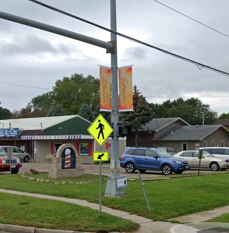 A street corner with a pedestrian crossing sign, a barber shop, cars parked in a lot, and promotional city banners.