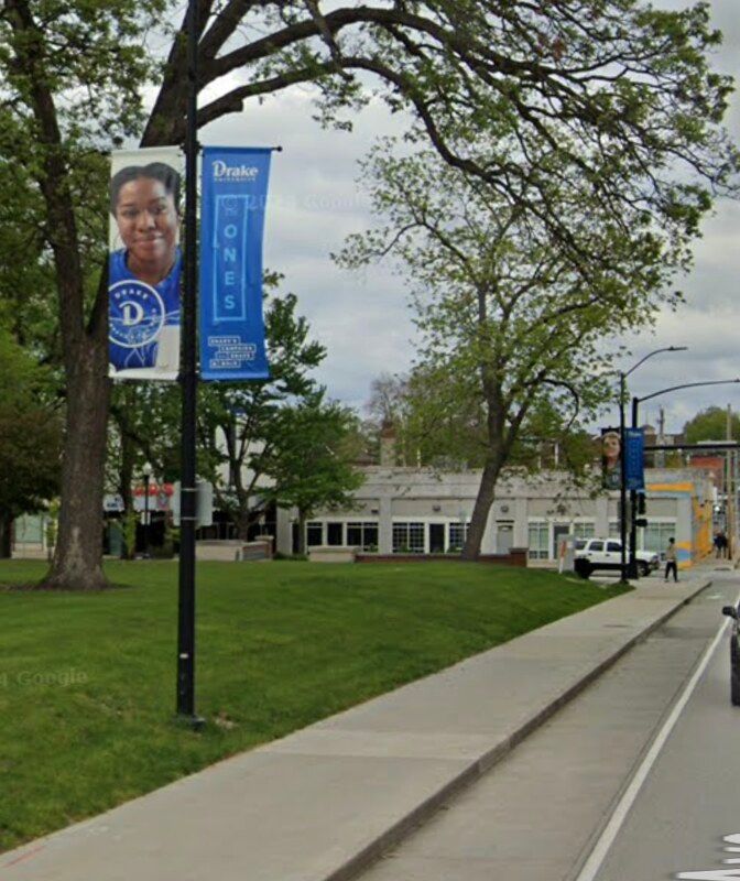 Drake University banners hang on a pole next to a sidewalk and green lawn near a campus building.