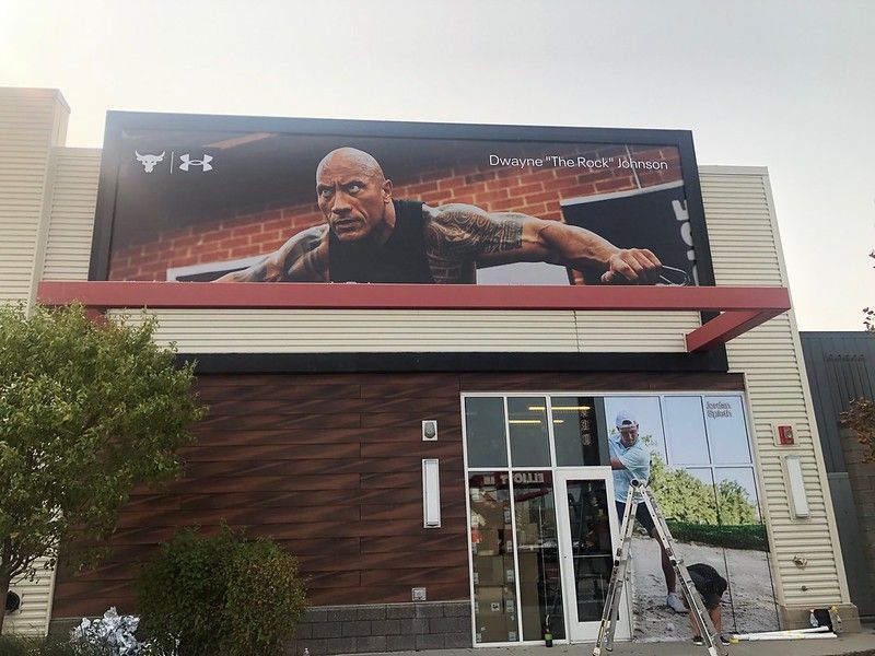 A large Under Armour advertisement featuring The Rock above a store entrance, with a person on a ladder working on windows.