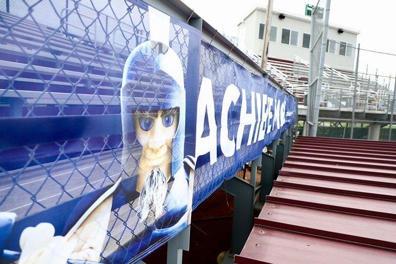 A blue banner featuring a mascot with a helmet hangs on a chain-link fence at a stadium with bleachers.
