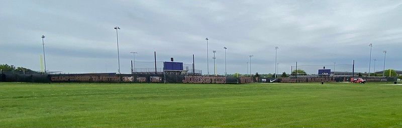 A wide, grassy field in front of a stadium with a tall perimeter fence and stadium lights under an overcast sky.