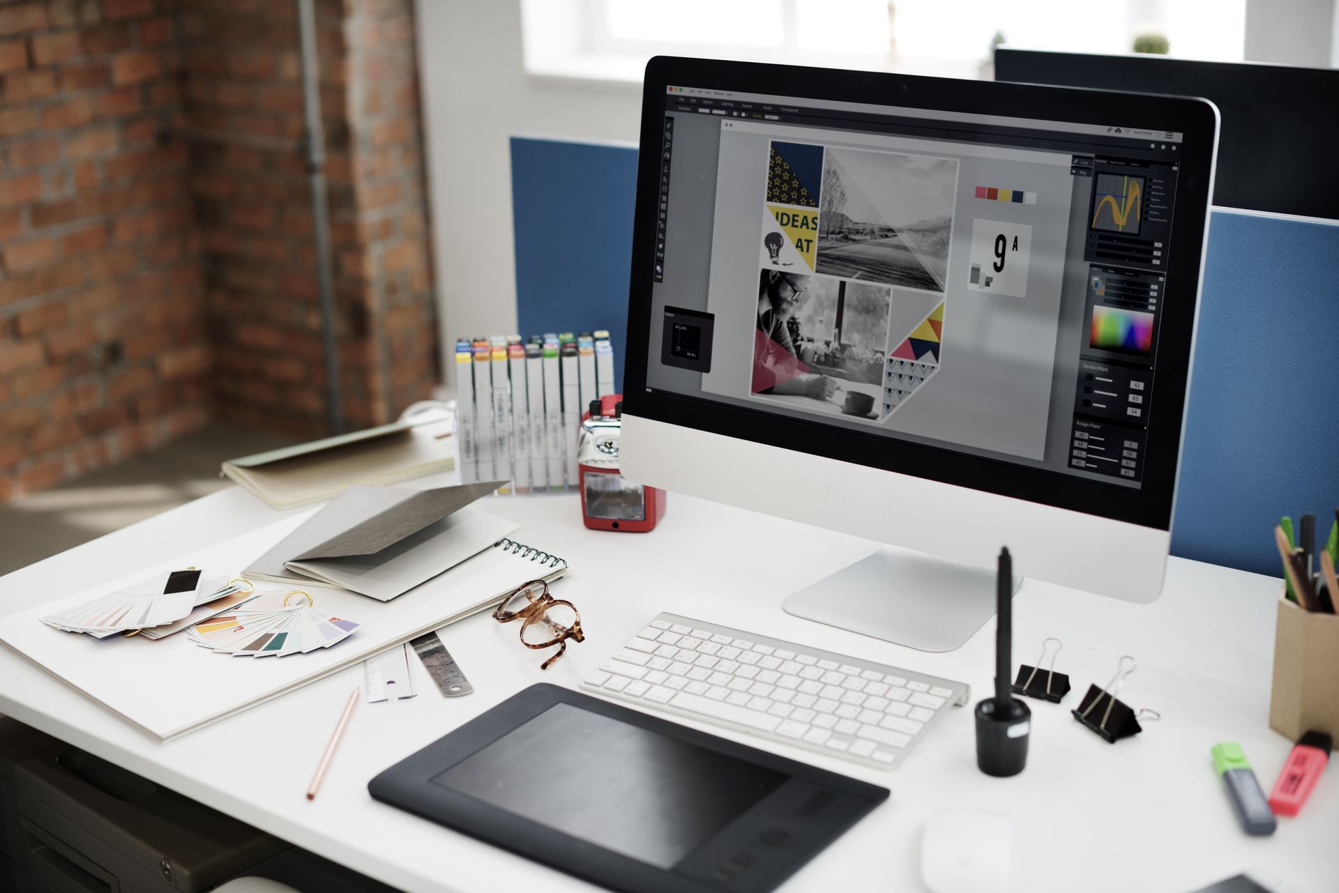 A designer’s white desk with a computer displaying graphics, a drawing tablet, markers, and supplies in an office space.