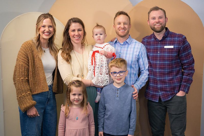 A smiling group of six poses in front of two large, beige arches; they wear casual clothing including sweaters and shirts.