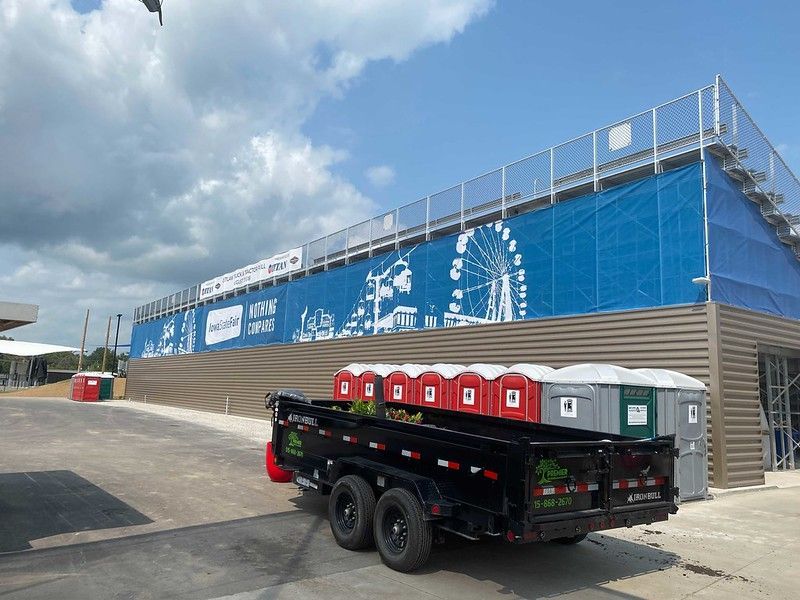 A black dump trailer parked in front of a stadium structure with a blue banner featuring fairground icons and port-a-potties.